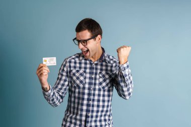 Mavi arka planda izole edilmiş kredi kartı gösteren mutlu genç adam.