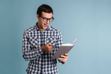 Yakışıklı, mutlu, genç bir adam mavi arka planda dikilirken notlar yazıyor.