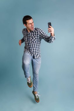 Mavi arka planda tek başına atlarken selfie çeken mutlu, genç bir adam.