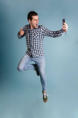 Mutlu, genç, sıradan bir adam mavi arka planda izole bir şekilde atlarken selfie çekiyor.