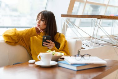 Çekici esmer kız kafeteryada kahve içerken cep telefonu kullanıyor.