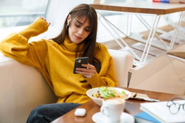 Kapalı kafede yemek yerken cep telefonu kullanan kulaklıklı hoş bir kız.
