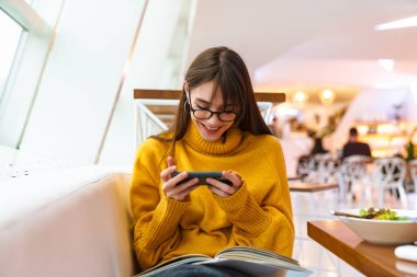 Gözlüklü, gülümseyen güzel kız kitap okuyor ve kapalı mekanda cep telefonu kullanıyor.