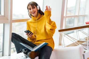 Evde kitap okurken kulaklıklı, el sallayan ve cep telefonu kullanan neşeli bir kız.