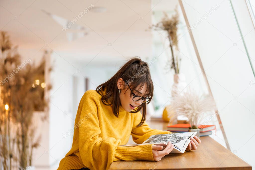 Charming beautiful smiling girl in eyeglasses reading book while sitting in cafe indoors