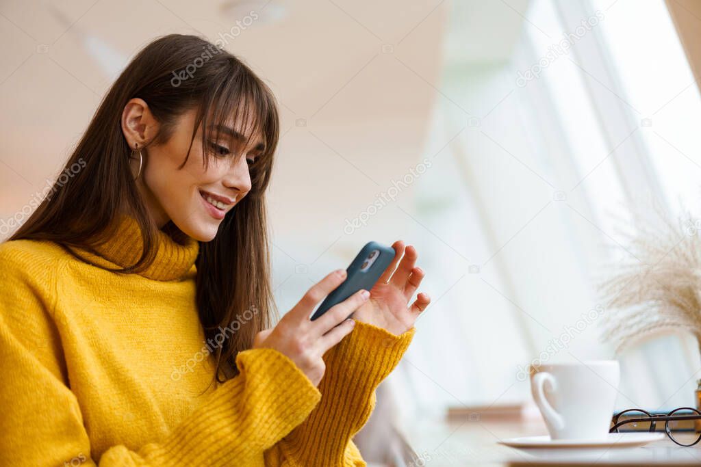Charming brunette happy girl using mobile phone while drinking coffee in cafe indoors