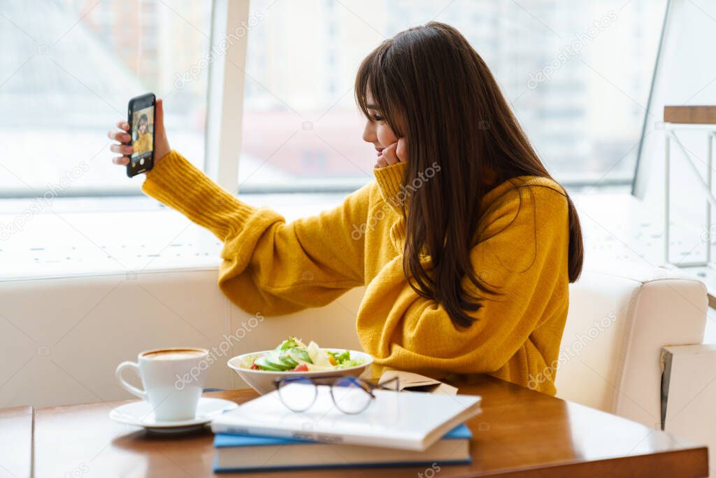 Charming happy girl taking selfie on mobile phone and having lunch in cafe indoors