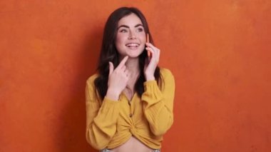 Stüdyoda turuncu arka planda tek başına duran genç bir kadın akıllı telefonuyla konuşuyor.