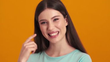 Mutlu genç bir kadın mükemmel gülüşünü işaret ediyor. Stüdyonun turuncu arka planında tek başına duruyor.