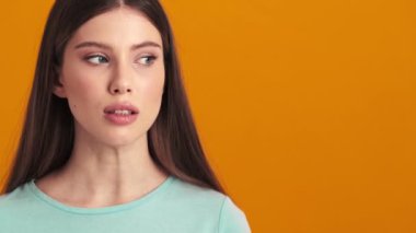 Güzel genç bir kadın yan tarafa bakıyor ve stüdyoda turuncu bir arka plan üzerinde izole bir şekilde duran gizli bir işaret veriyor.