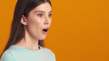 Şaşırmış genç bir kadın yan tarafa bakıyor ve gizli bir jest yapıyor. Stüdyoda turuncu bir arka planda tek başına duruyor.