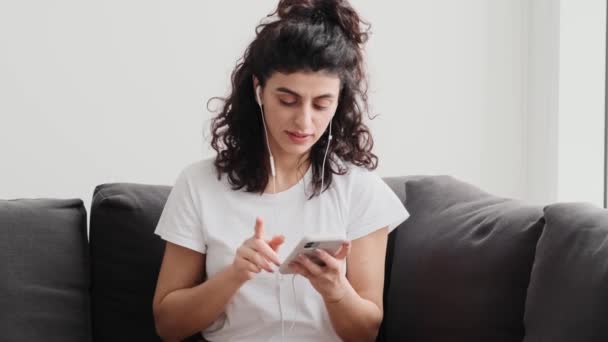 Soustředěná Mladá Žena Sluchátky Používá Svůj Smartphone Zatímco Sedí Doma — Stock video