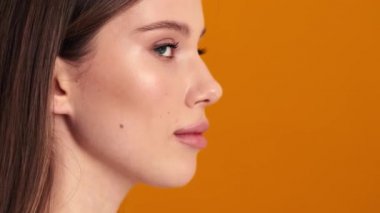 Gülümseyen genç bir kadının yakın çekim görüntüsü başını kameraya çeviriyor sonra da stüdyonun turuncu arka planından izole edilmiş bir şekilde yan tarafa bakıyor.