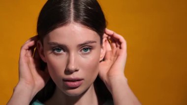 Stüdyoda turuncu bir arka planda tek başına duran genç ve seksi bir kadının yüzüne dokunurken yakın çekim görüntüsü kameraya yansıyor.
