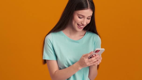 一位穿着基本T恤 面带微笑的年轻女子正在工作室里用她的智能手机站在橙色的背景下 — 图库视频影像