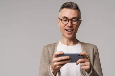 Gri arka planda izole bir şekilde cep telefonuyla oyun oynayan heyecanlı ve zeki görünümlü bir adamın portresi