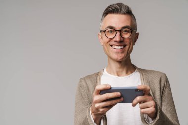 Gri arka planda izole bir şekilde cep telefonuyla oyun oynayan heyecanlı ve zeki görünümlü bir adamın portresi