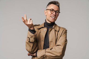 Yakışıklı, güler yüzlü, şık bir ceket giyen, gri arka planda yalnız poz veren adam.