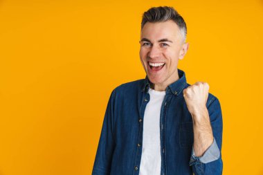 Heyecanlı yakışıklı adam sarı arka planda izole edilmiş kazanan jestler yapıyor.