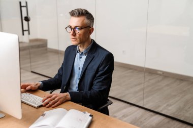 Ofiste otururken bilgisayarla çalışan odaklanmış gri adam.