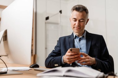 Ofiste bilgisayarla çalışırken cep telefonu kullanan memnun gri adam.