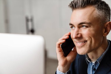 Ofiste bilgisayarla çalışırken cep telefonuyla konuşan gülümseyen gri adam.