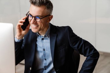 Ofiste bilgisayarla çalışırken cep telefonuyla konuşan gülümseyen gri adam.