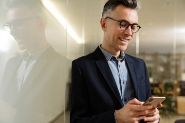 Ofiste duvara yaslanırken cep telefonu kullanan gülümseyen gri adam.