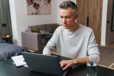 Ciddi gri adam evde oturma odasında otururken dizüstü bilgisayarla çalışıyor.