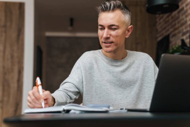 Memnuniyet verici gri adam notlar yazıyor ve evde otururken bilgisayarla çalışıyor.