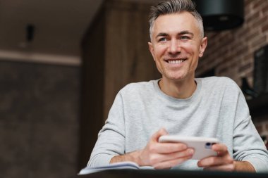 Evde çalışırken gülümseyen ve cep telefonu kullanan neşeli yakışıklı bir adam.