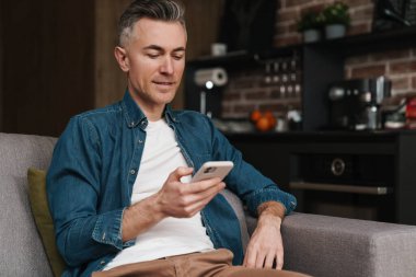 Yakışıklı orta yaşlı adam evde akıllı telefon kullanıyor, kanepede oturuyor.