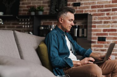 Evde kanepede otururken dizüstü bilgisayar kullanan yakışıklı bir adam.