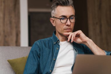 Gözlüklü yakışıklı adam evde kanepede otururken dizüstü bilgisayar kullanıyor.