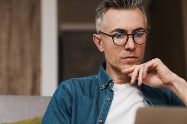 Gözlüklü yakışıklı adam evde kanepede otururken dizüstü bilgisayar kullanıyor.