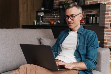 Gözlüklü gülümseyen yakışıklı adam evdeki kanepede otururken dizüstü bilgisayar kullanıyor.