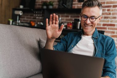 Evdeki kanepede otururken el sallayan ve dizüstü bilgisayar kullanan neşeli yakışıklı adam.