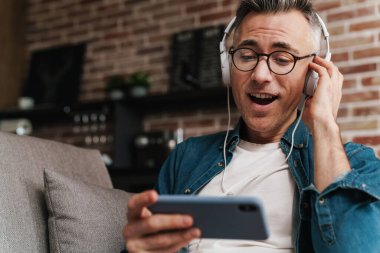 Evde koltukta otururken cep telefonu kullanan kulaklıklı heyecanlı adam.