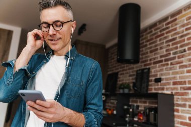 Gözlüklü, kulaklık ve cep telefonu kullanan neşeli, beyaz saçlı bir adam.