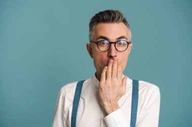 Gözlüklü, ağzı kapalı, mavi arka planda mahsur kalmış gri saçlı, şaşırmış bir adam.