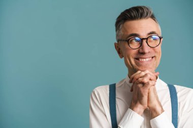 Gözlüklü mutlu gri saçlı adam mavi arka planda izole bir şekilde poz veriyor ve yan bakıyor.