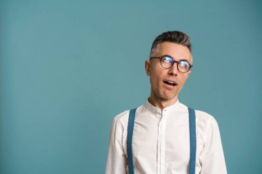 Kafası karışmış gözlüklü gri saçlı adam yüzünü buruşturuyor ve yukarı bakıyor mavi arka planda soyutlanmış.