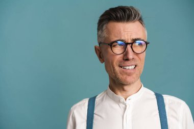 Kafası karışmış gözlüklü gri saçlı adam yüzünü buruşturuyor ve mavi arka planda yalnız başına bakıyor.