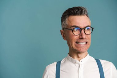 Gözlüklü hoşnutsuz gri saçlı adam yüzünü buruşturuyor ve mavi arka planda yalnız başına bakıyor.