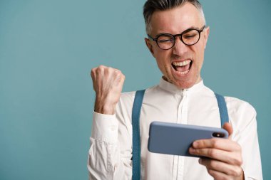 Heyecanlı adam mavi arka planda izole edilmiş bir cep telefonuyla çevrimiçi oyun oynarken kazanan hareketi yapıyor