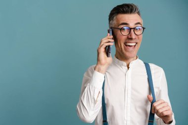 Heyecanlı, gözlüklü, mavi arka planda izole bir şekilde cep telefonuyla konuşan gri saçlı adam.