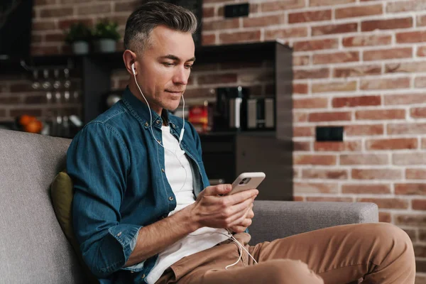 Focused handsome man in earphones using mobile phone while sitting on couch at home