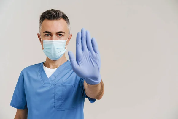 Serious medical doctor in face mask and gloves showing stop gesture isolated over white wall