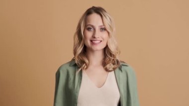 Gündelik giysiler giyen gülümseyen genç bir kadın, stüdyoda bej arka planda tek başına dururken baş parmak hareketi gösteriyor.