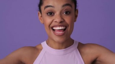 Günlük giysiler giyen neşeli genç bir kadın, elleri ile yüzünü kapatırken kameraya poz veriyor. Stüdyonun menekşe arka planında yalnız duruyor.